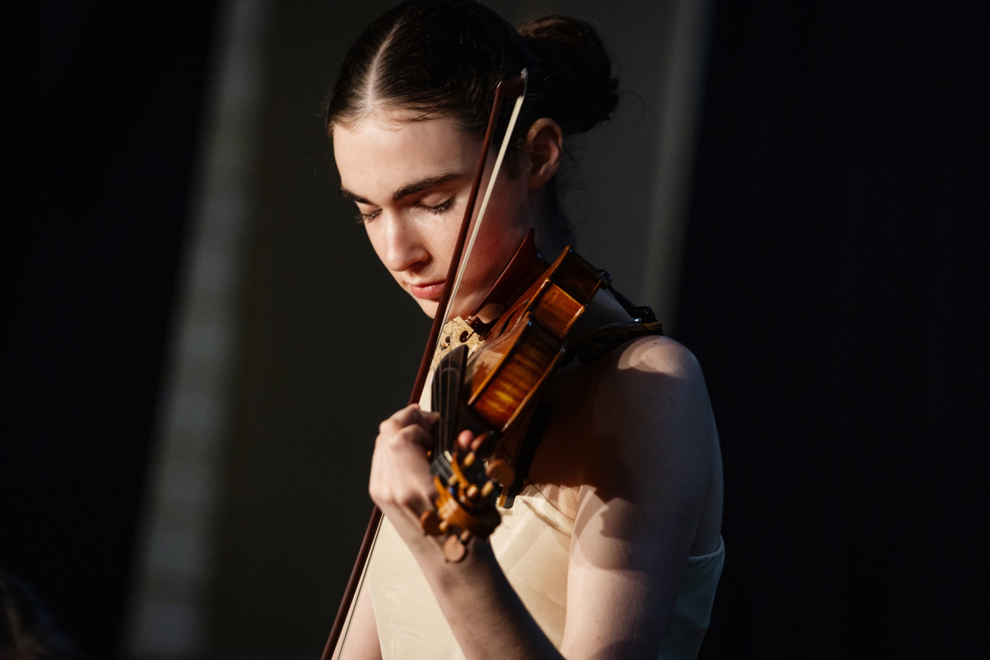 A violinist performs a solo, eyes closed in concentration, lit from the side on a dark stage.