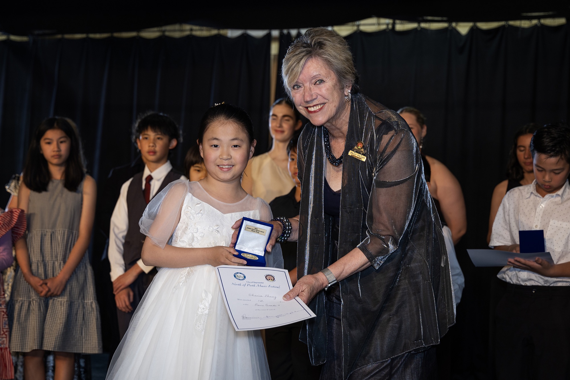 A young performer in a white dress receives her certificate on stage.