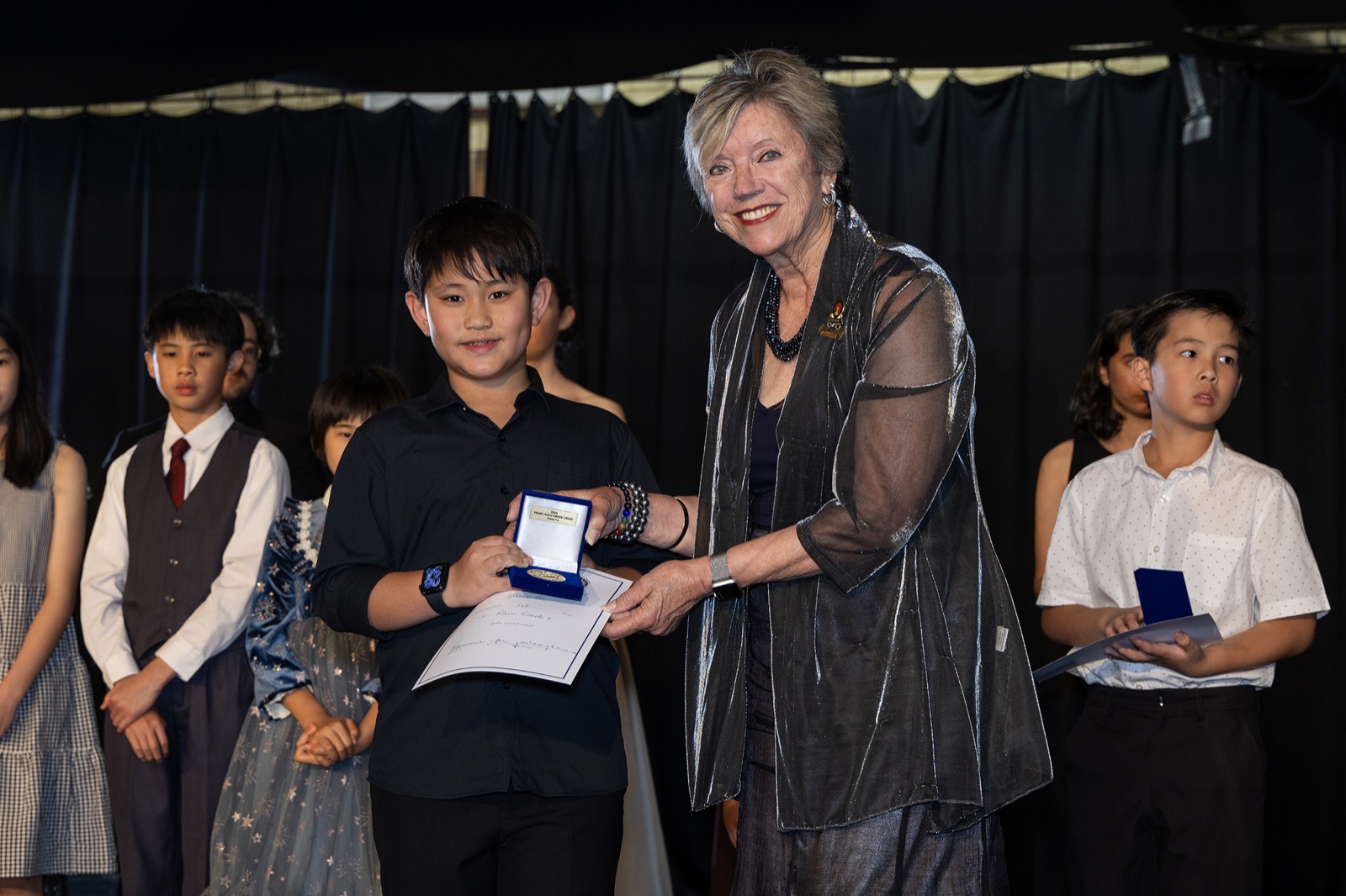 An adjudicator presents a medallion and certificate to a junior entrant, fellow entrants waiting alongside.