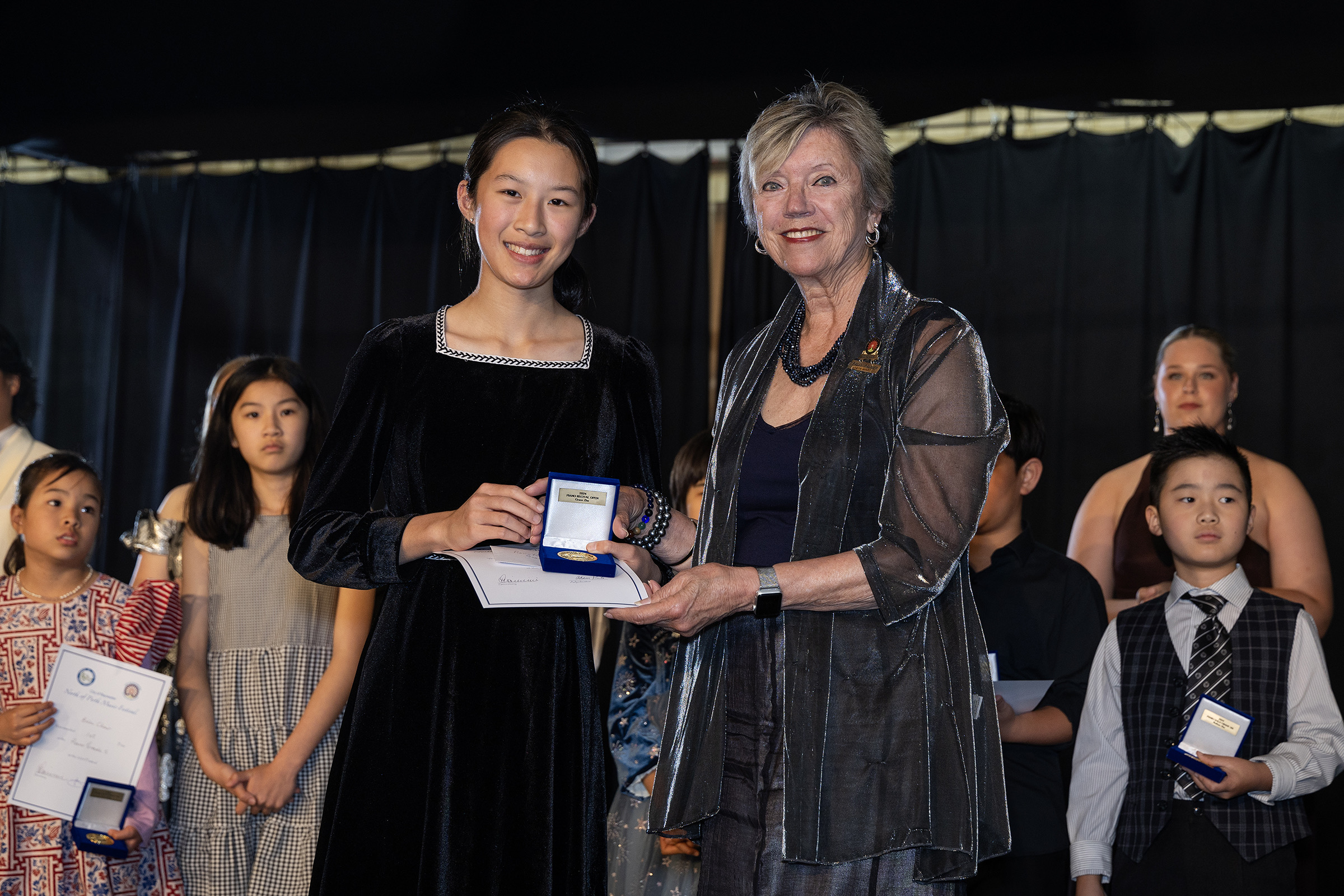 A young winner in a black velvet dress receives her award from a presenter, fellow entrants looking on.